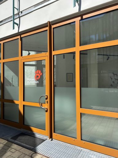 Braune Glastür mit rotem Logo, umgeben von großen Fensterflächen.